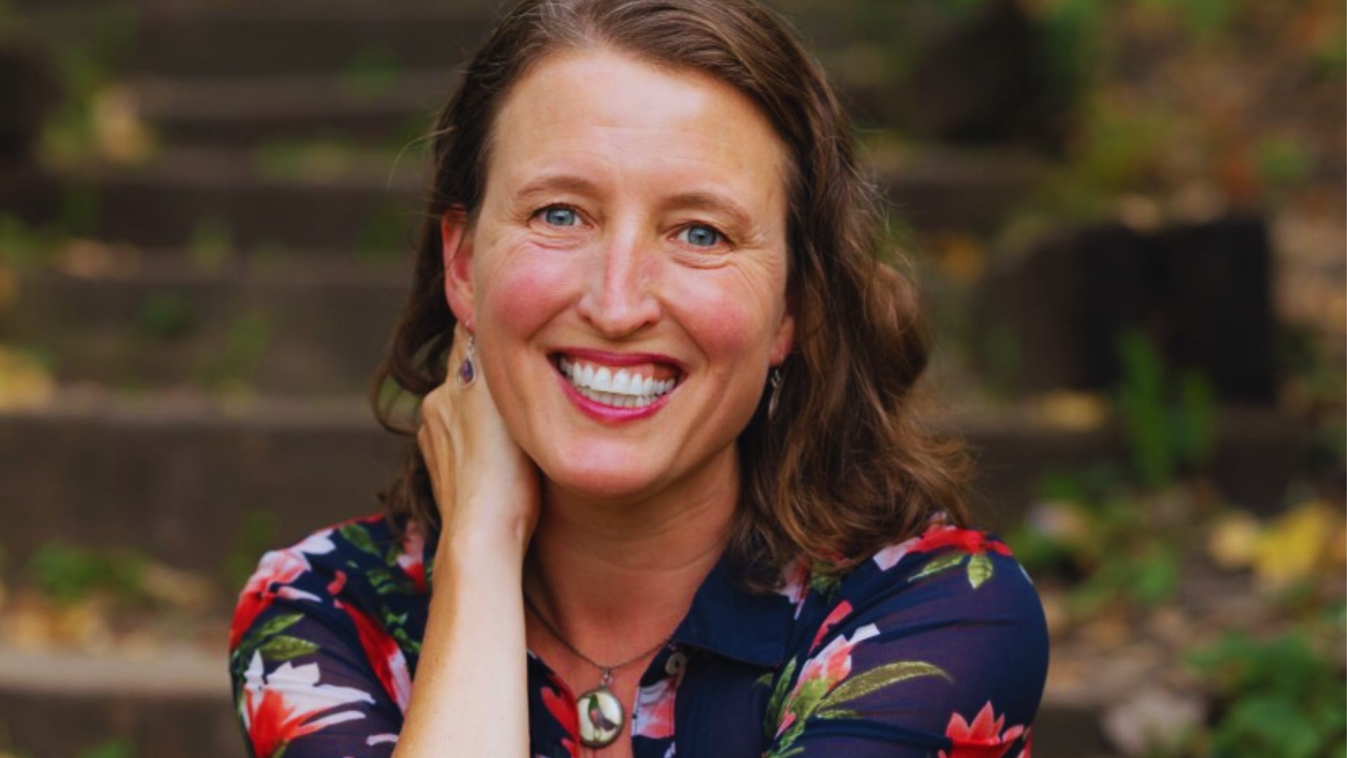 A smiling Kelly Barnhill sits on steps, radiating joy and contentment in a relaxed outdoor environment.