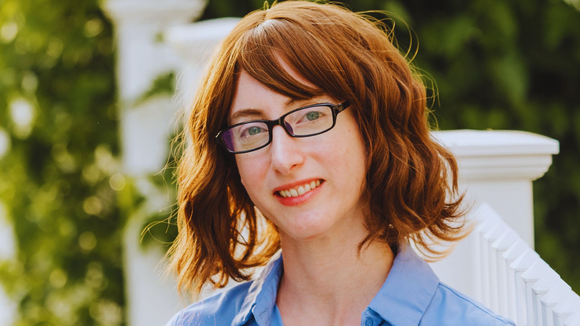 A woman in a blue shirt and glasses, standing with a friendly expression against a neutral background.