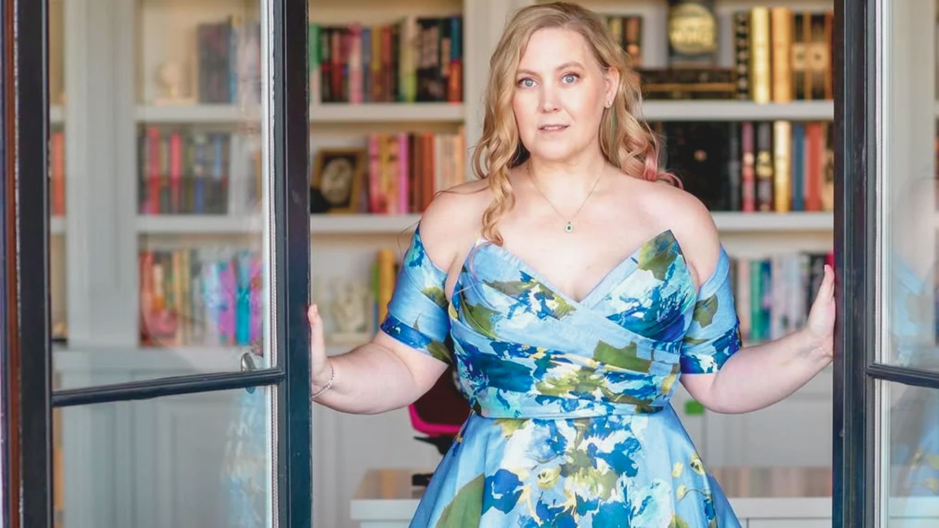 A woman wearing a blue floral dress poses in front of a bookcase lined with various books.