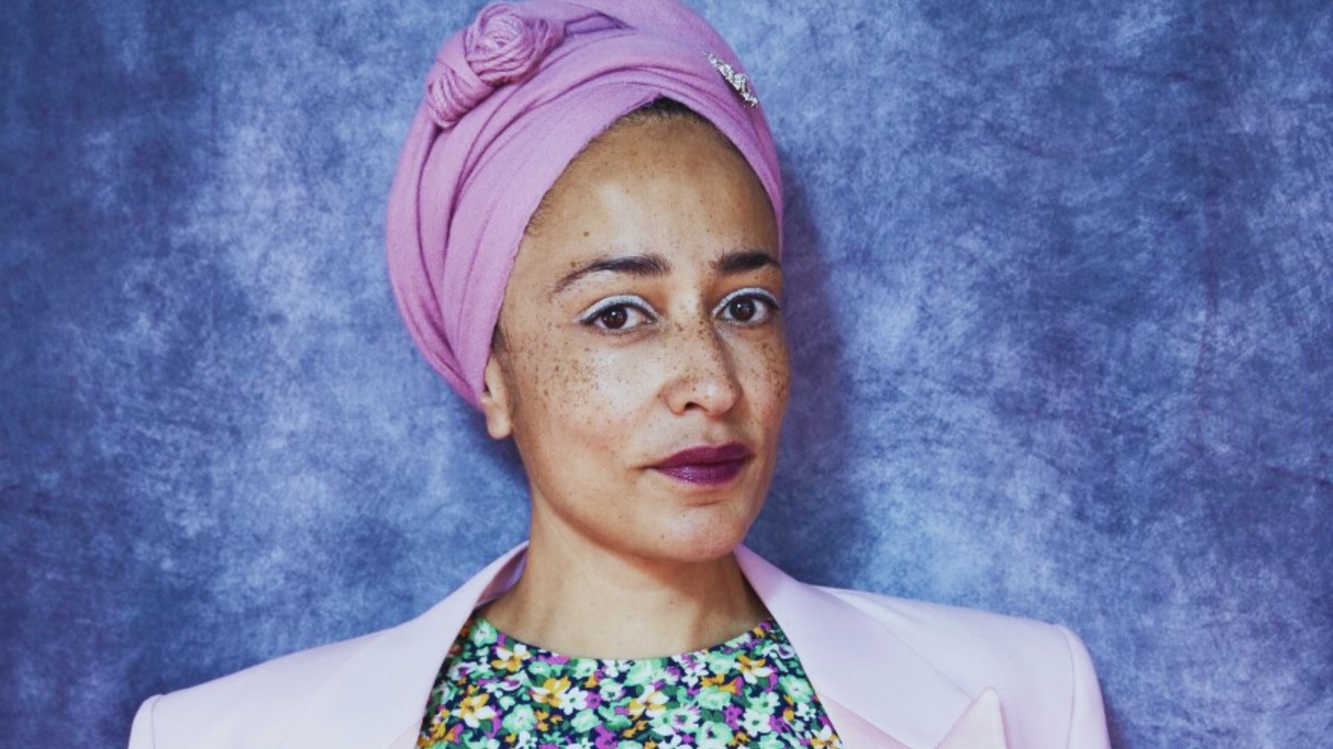 A woman dressed in a pink headscarf and pink jacket, standing with her arms crossed and a friendly smile.