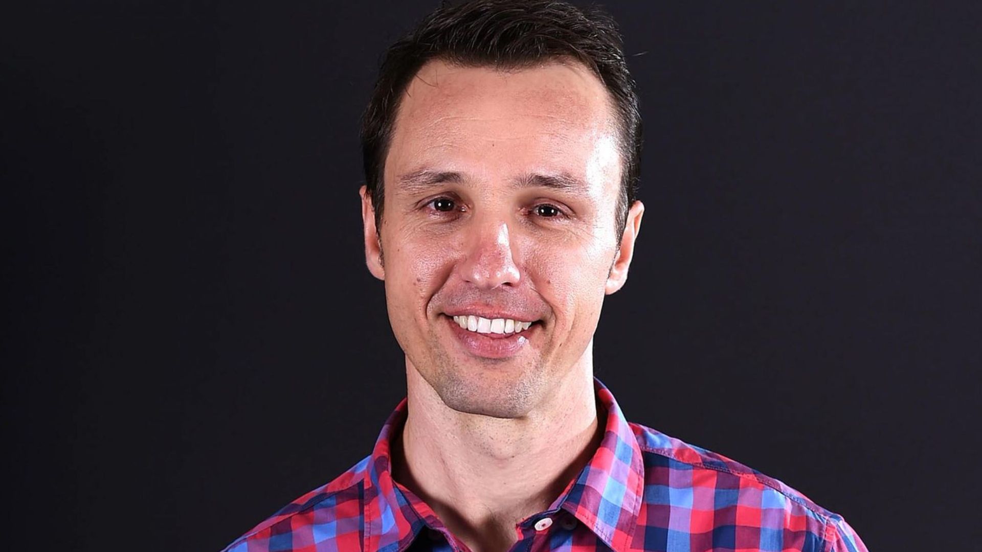 A man wearing a red and blue checkered shirt smiles warmly at the camera.