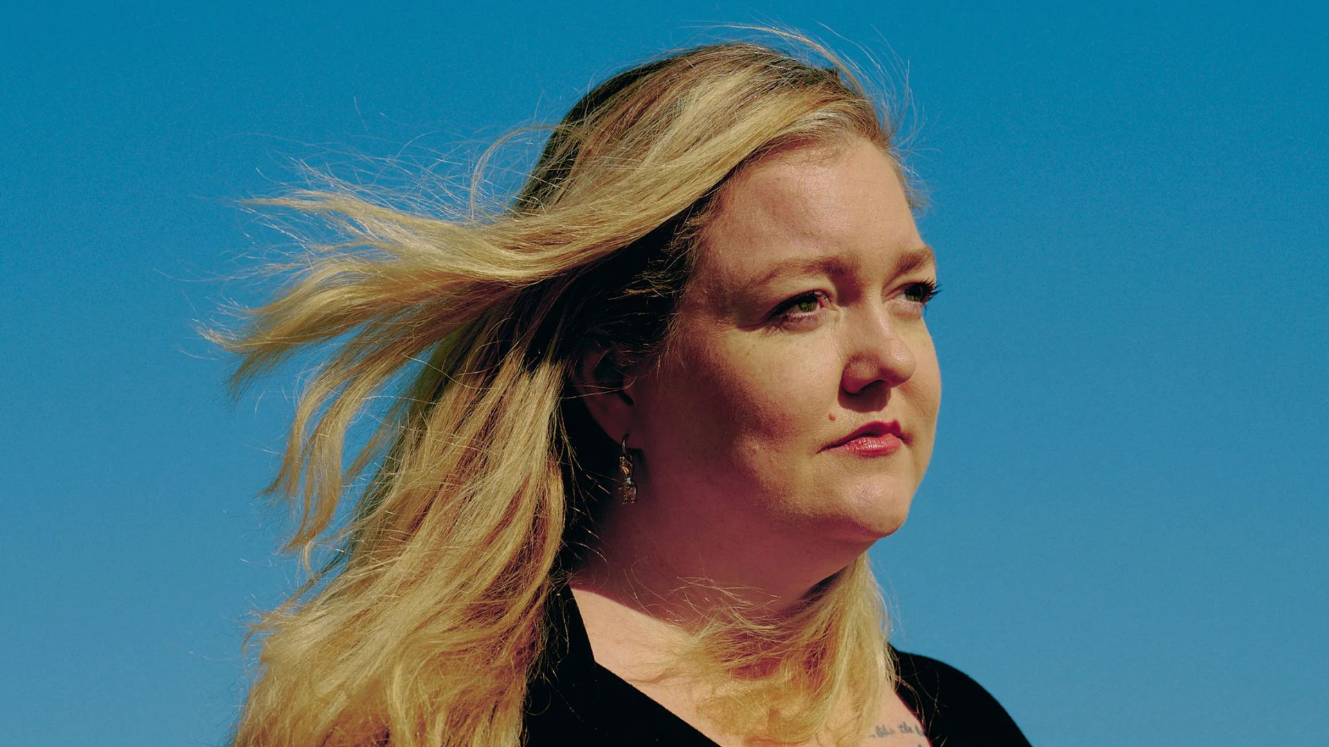 A woman with long blonde hair stands against a clear blue sky, radiating confidence and serenity.