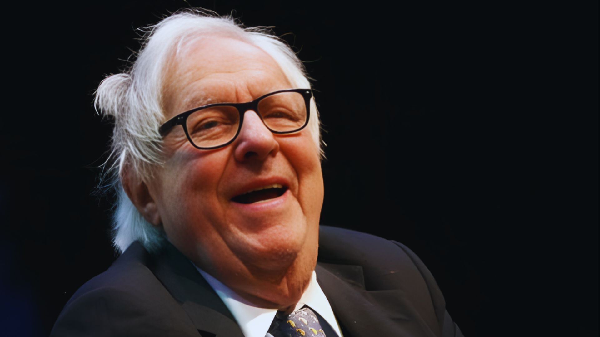 A smiling older man wearing glasses and a suit, exuding a friendly demeanor.