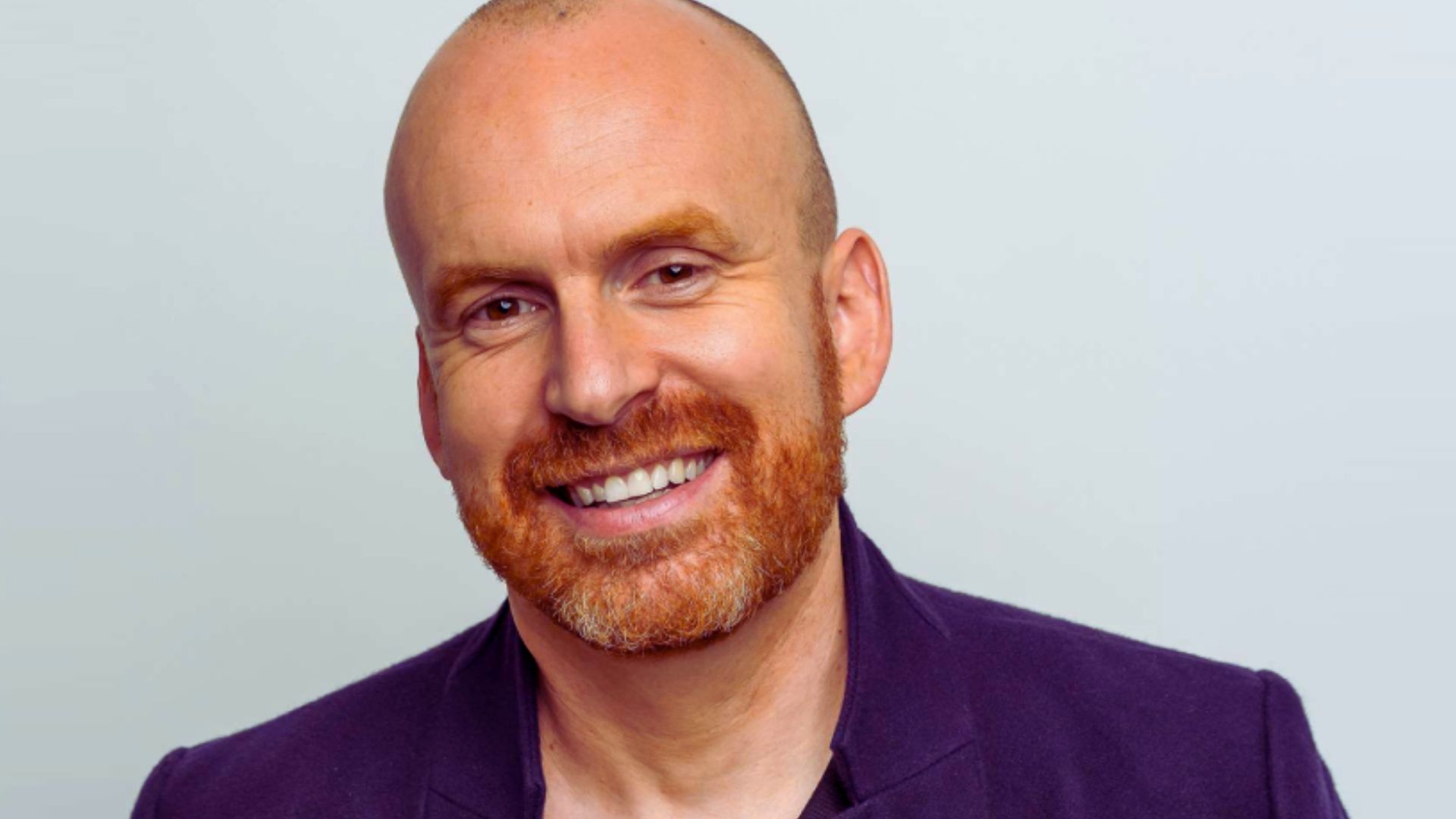 Matt Haig, a bald man with a beard, smiles cheerfully at the camera.