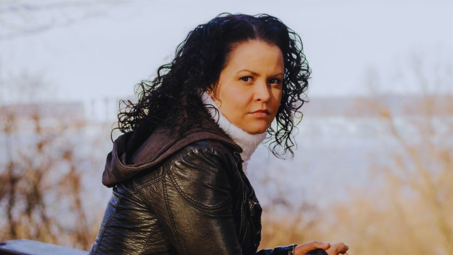 Jeanine Cummins, a woman with curly hair in a black jacket, sits on a bench, contemplating her journey as a writer.