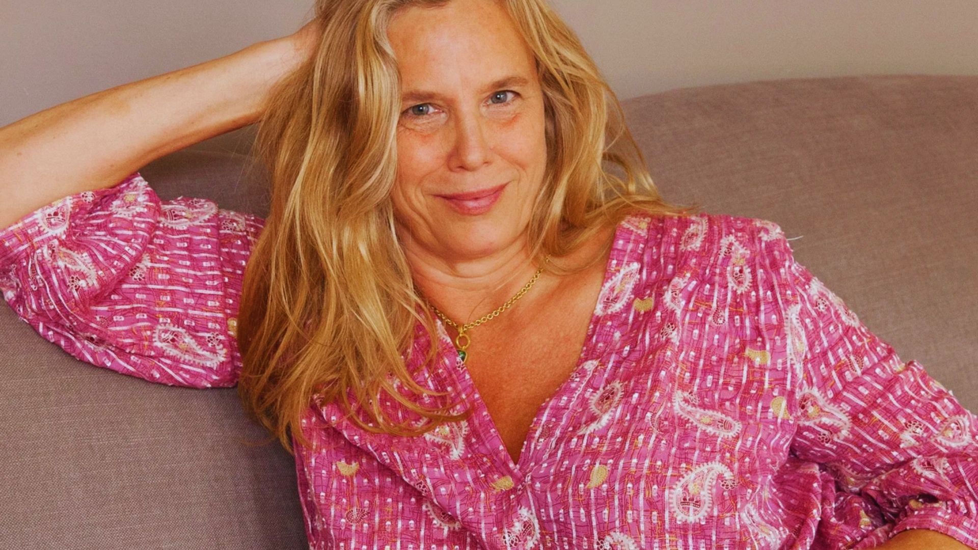 A woman with long blonde hair smiles softly while reclining on a gray sofa. She's wearing a pink patterned blouse, exuding a relaxed and cheerful vibe.