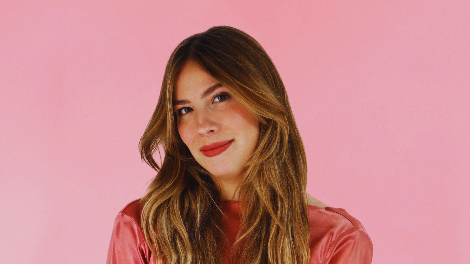 Portrait of a smiling person with long brown hair wearing a pink shirt. The background is solid pink, creating a cheerful and warm atmosphere.