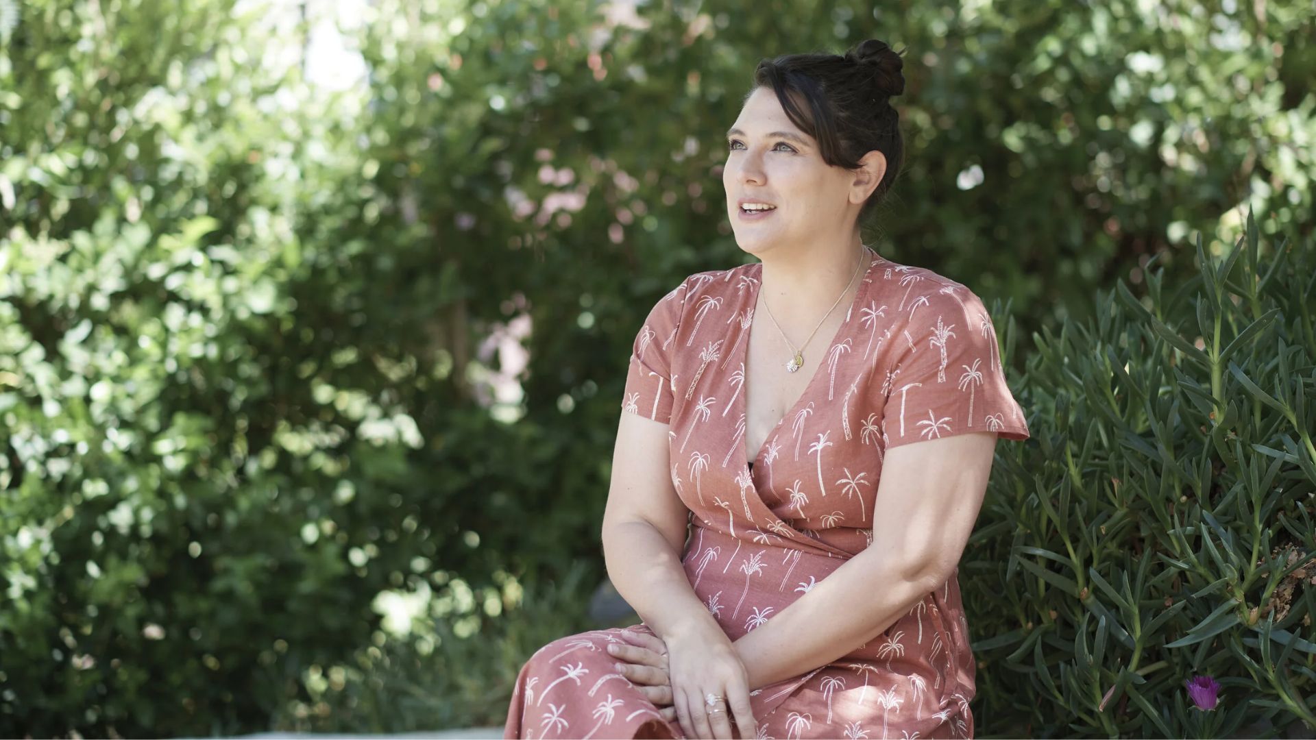 A woman wearing a dress is seated on a bench, appearing relaxed and contemplative.