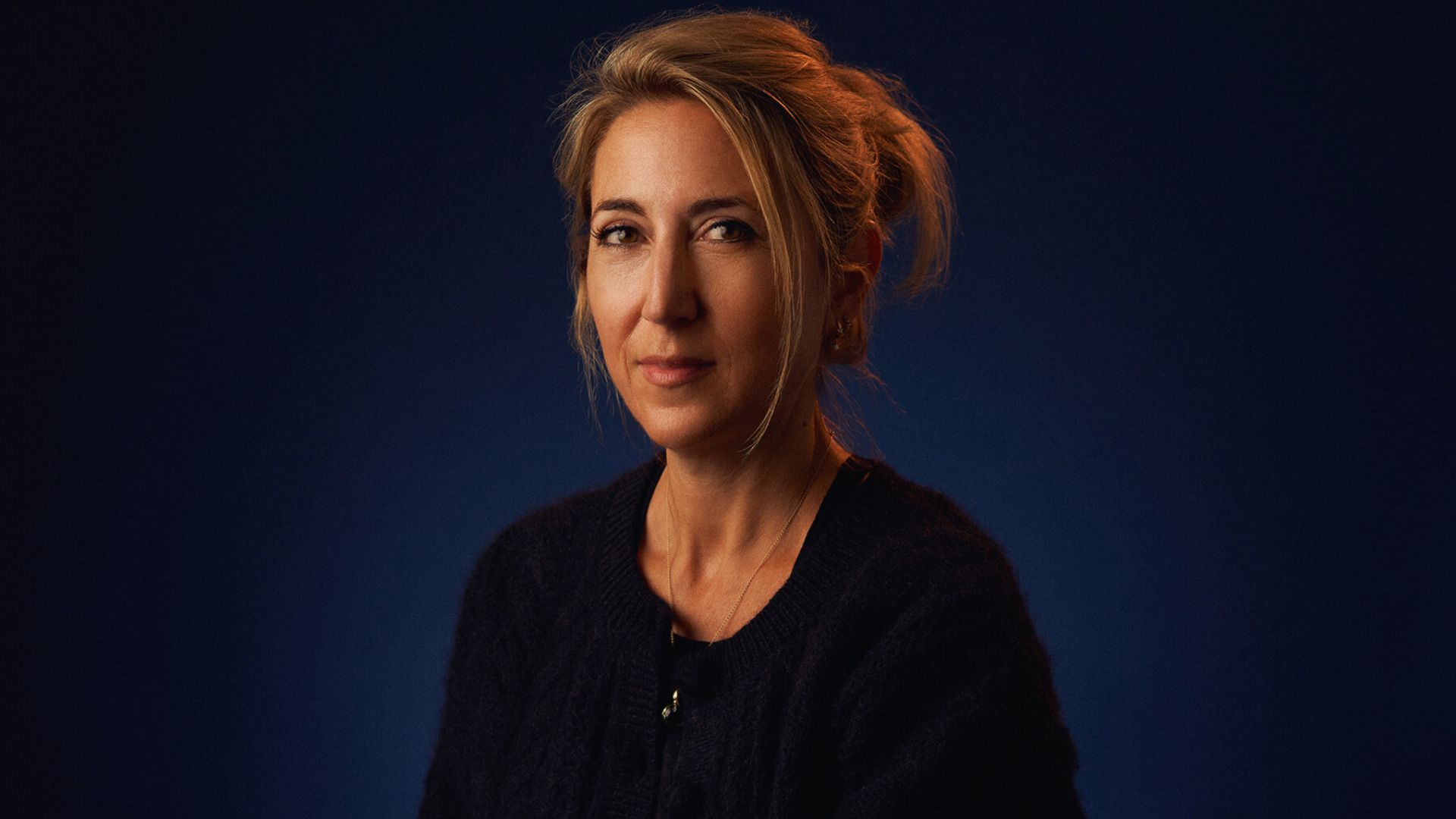 A woman dressed in a black sweater sits before a blue backdrop, her gaze directed towards the camera.