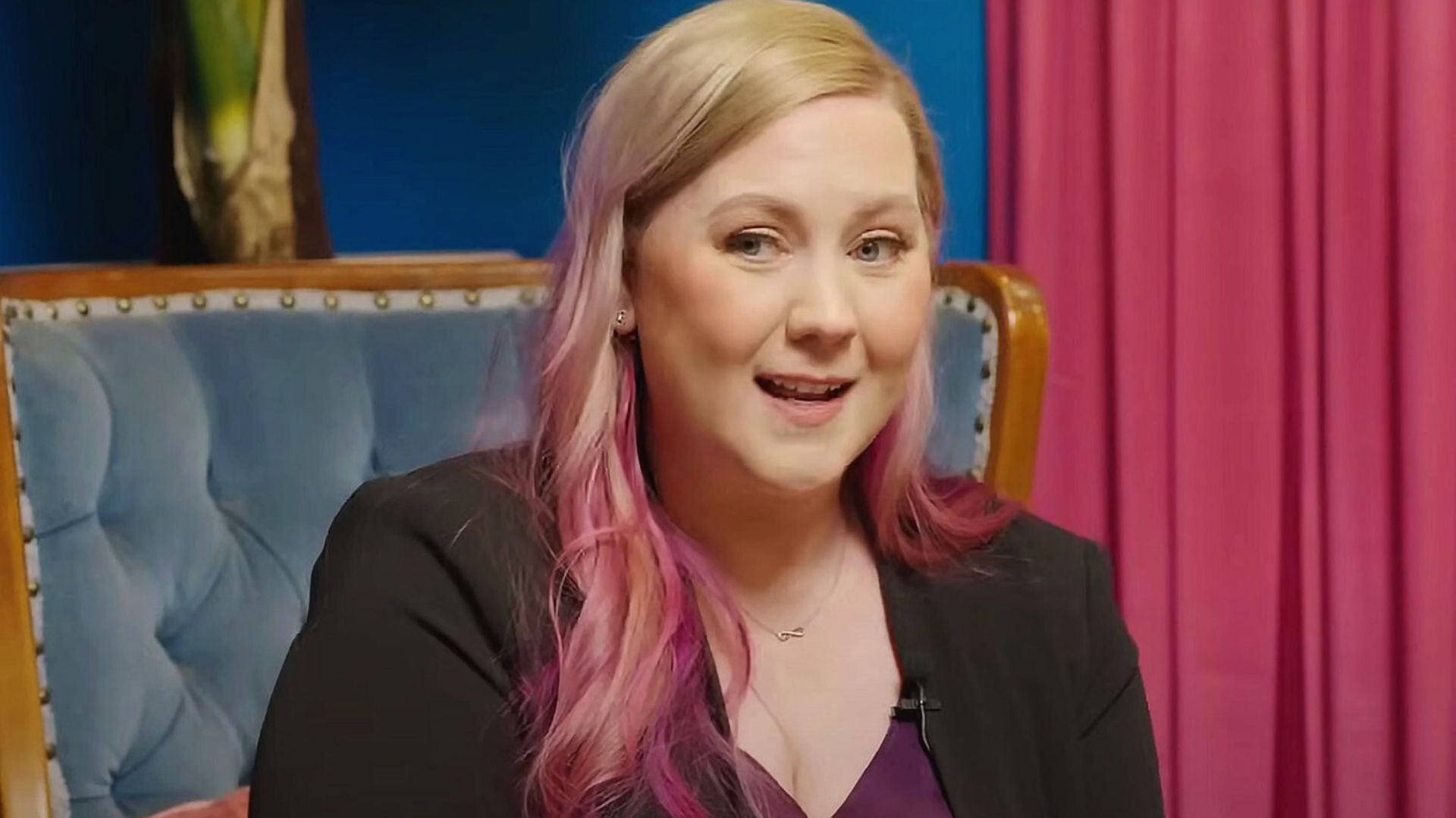 A woman with pink hair is sitting comfortably on a couch, looking relaxed and engaged in her surroundings.