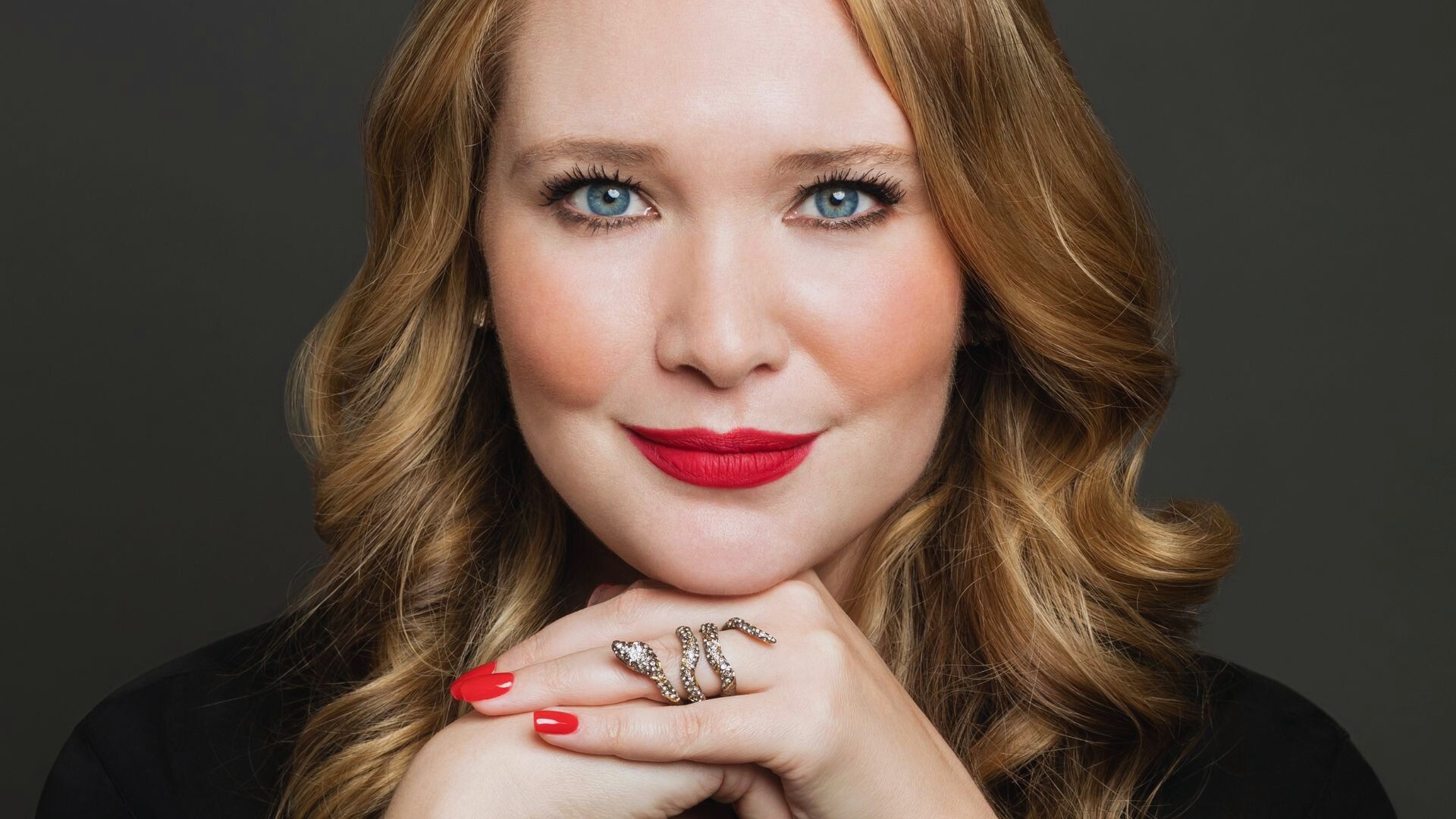 A woman displays her hand with red nails and a decorative ring, emphasizing her stylish manicure and jewelry.