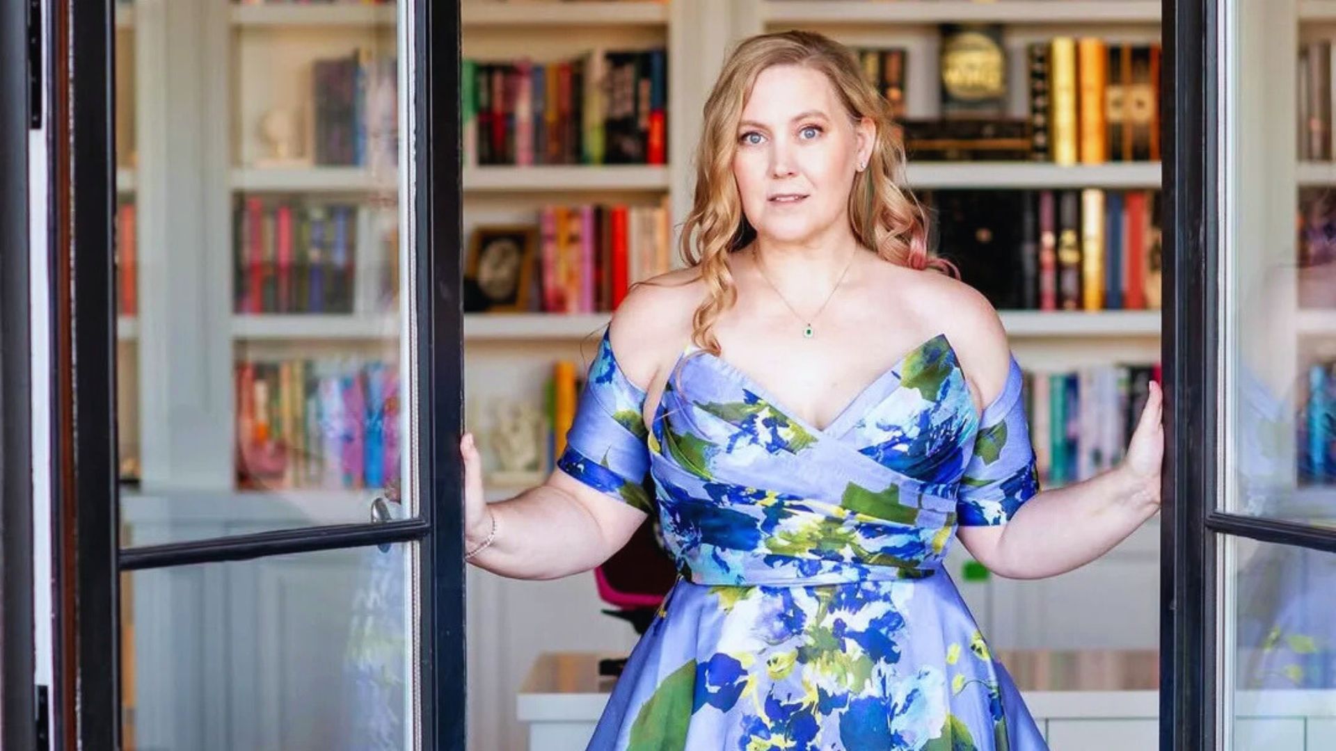 A woman in a blue floral dress stands in front of a bookcase filled with books.
