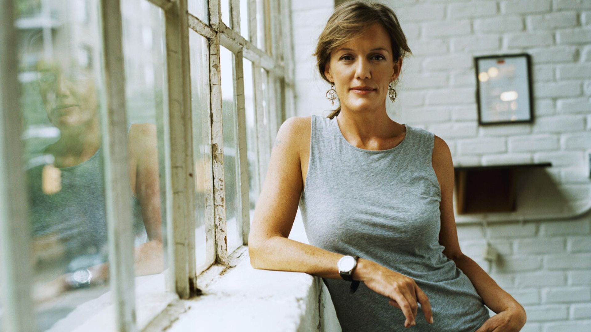 A woman in a gray tank top leans against a window, gazing thoughtfully outside.