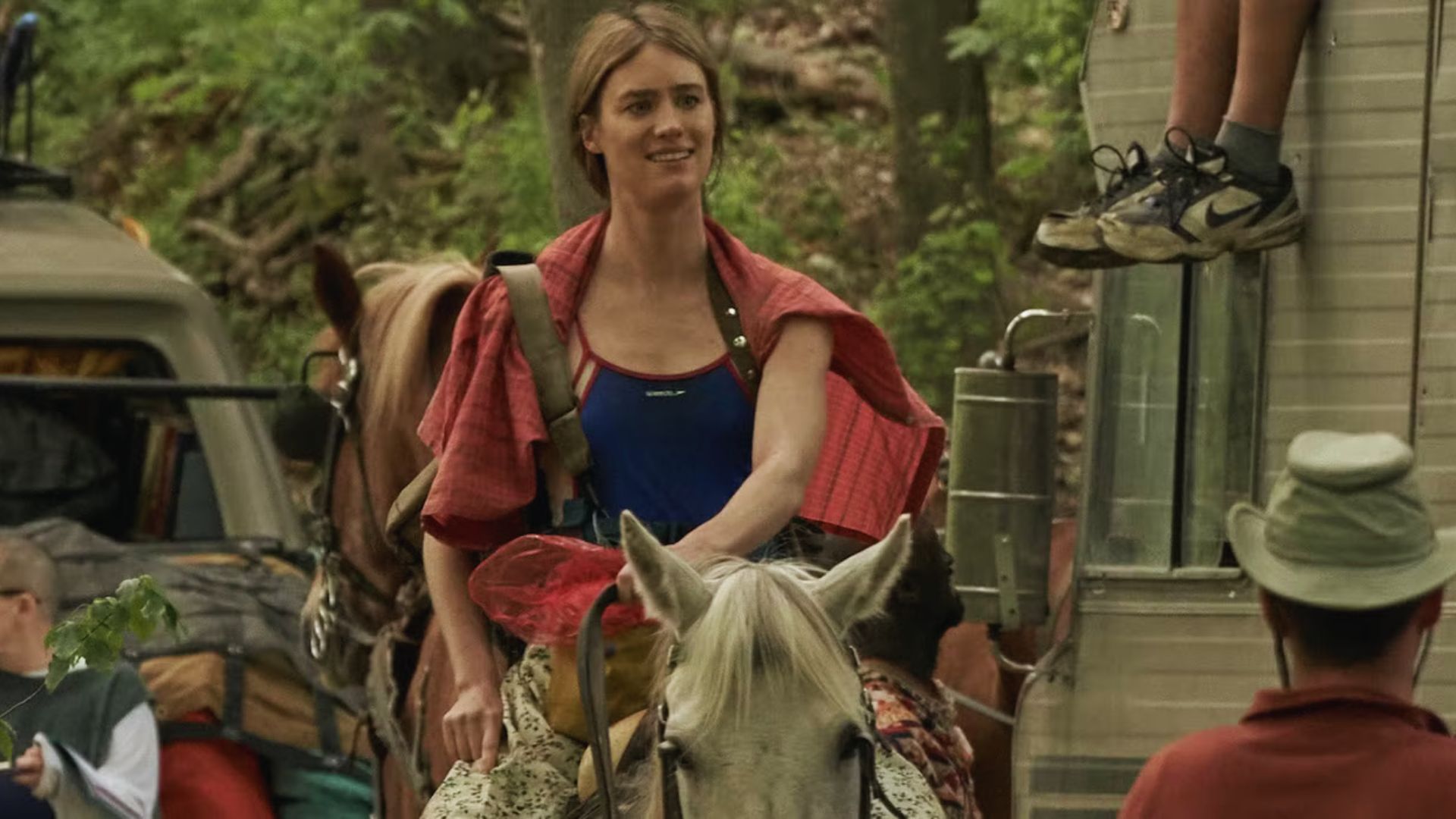 A woman on horseback stands in front of a trailer, surrounded by green grass.