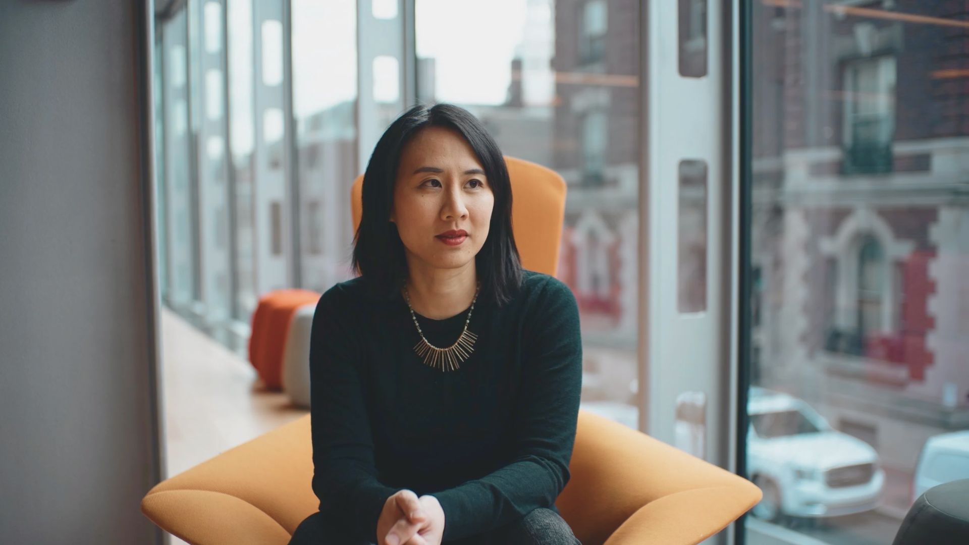 A woman sitting in an orange chair, with a window providing natural light in the background.