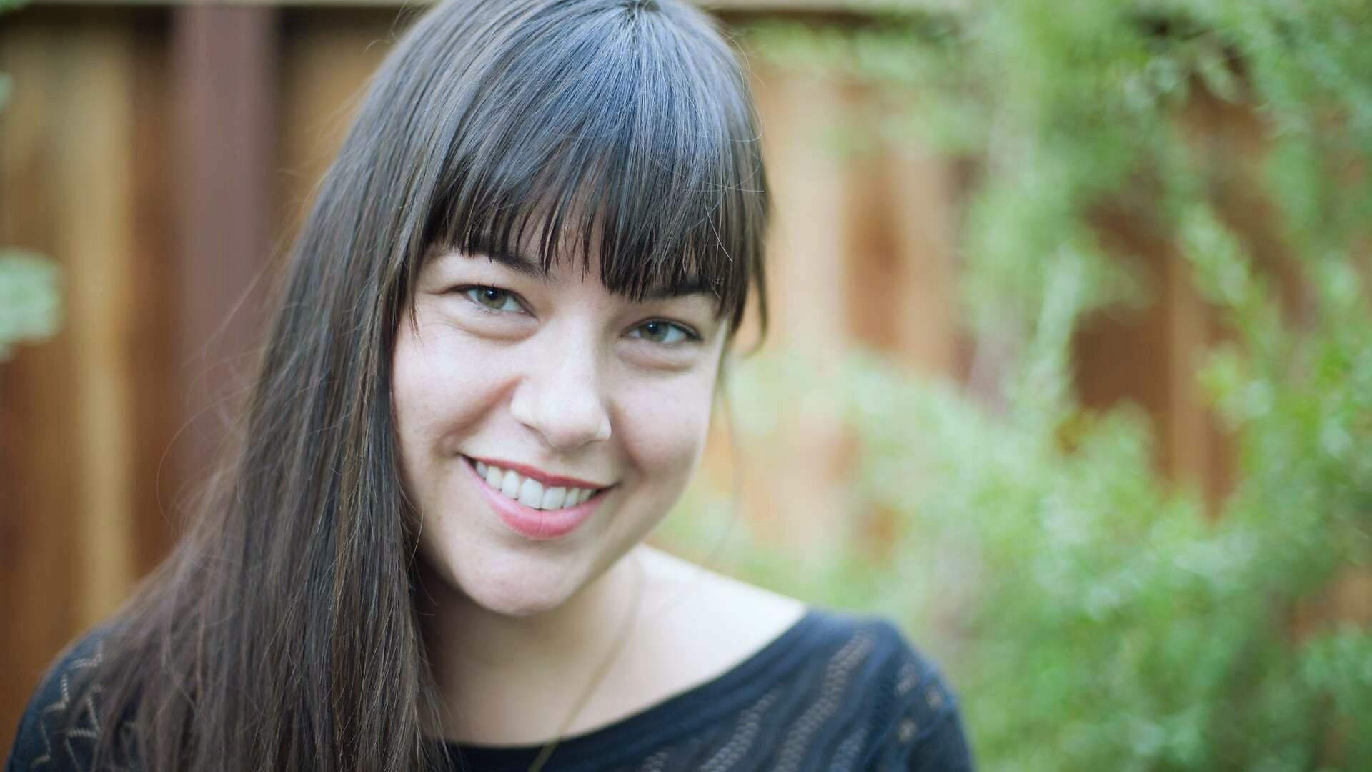 A woman with long hair smiles brightly in front of a wooden fence, radiating joy and warmth.
