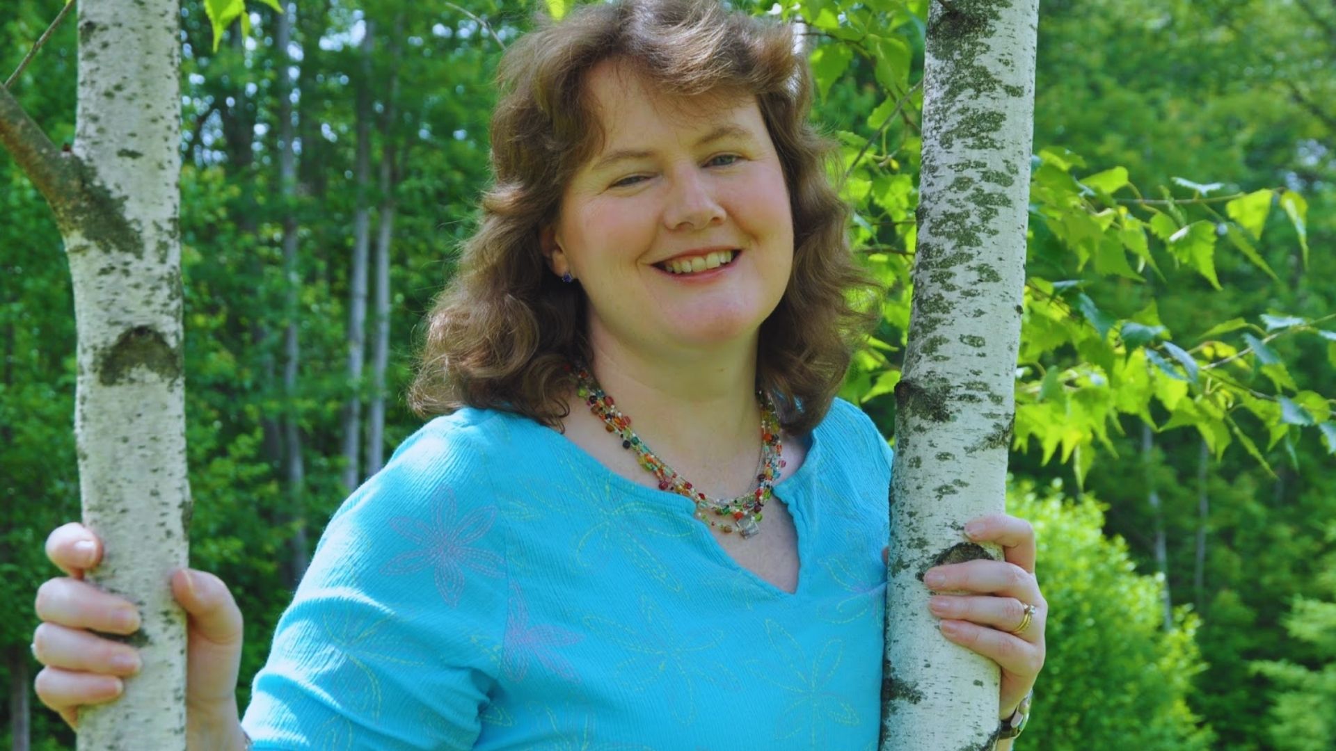 A woman stands beside a birch tree, smiling and enjoying the natural surroundings in a sunny outdoor setting.