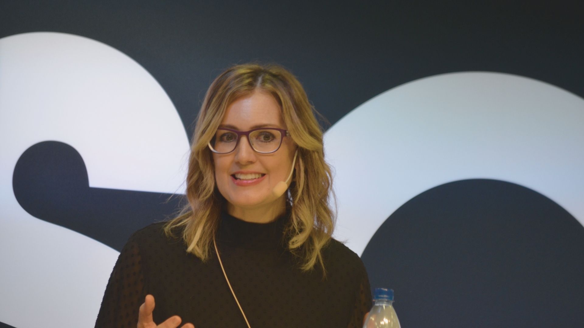A woman wearing glasses stands in front of a logo, smiling confidently at the camera.