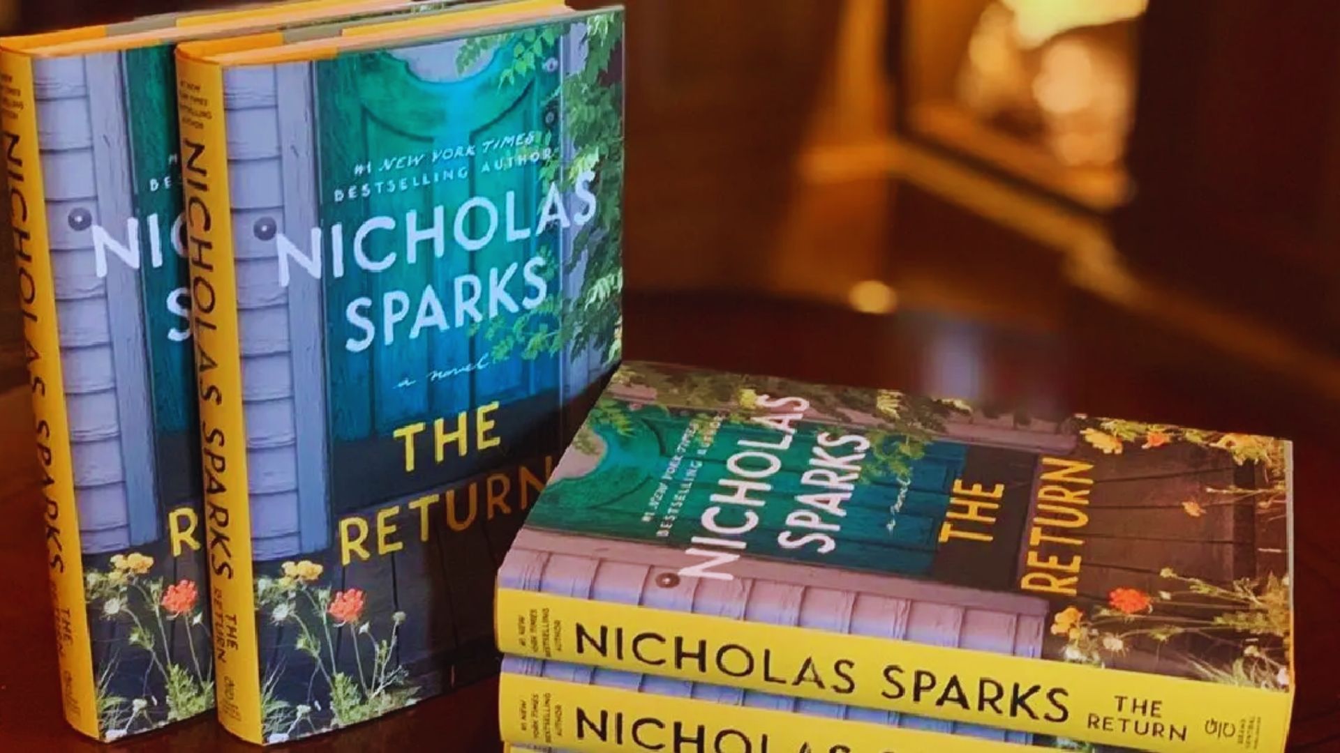 Four copies of the book "The Return" by Nicholas Sparks are displayed on a wooden table. The cover shows a green garden door with flowers.