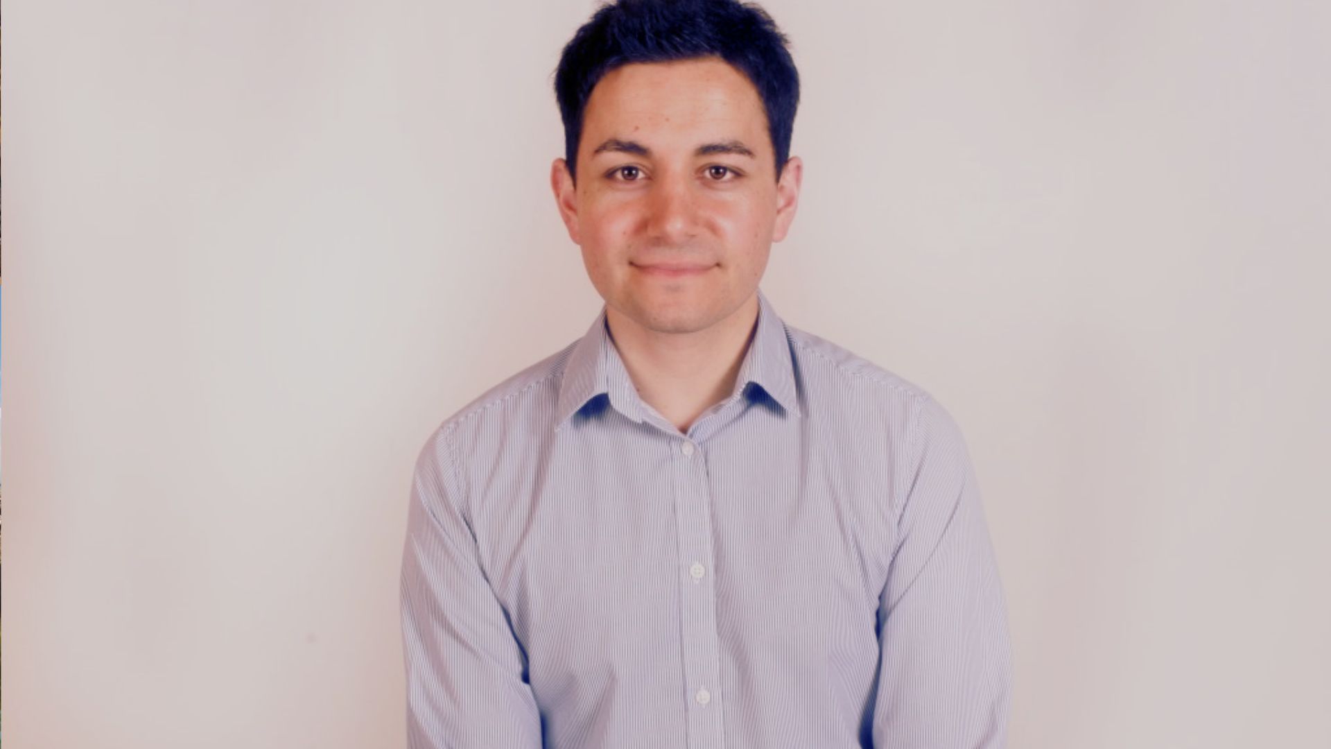 A man in a blue shirt and tie stands confidently in front of a plain white wall.
