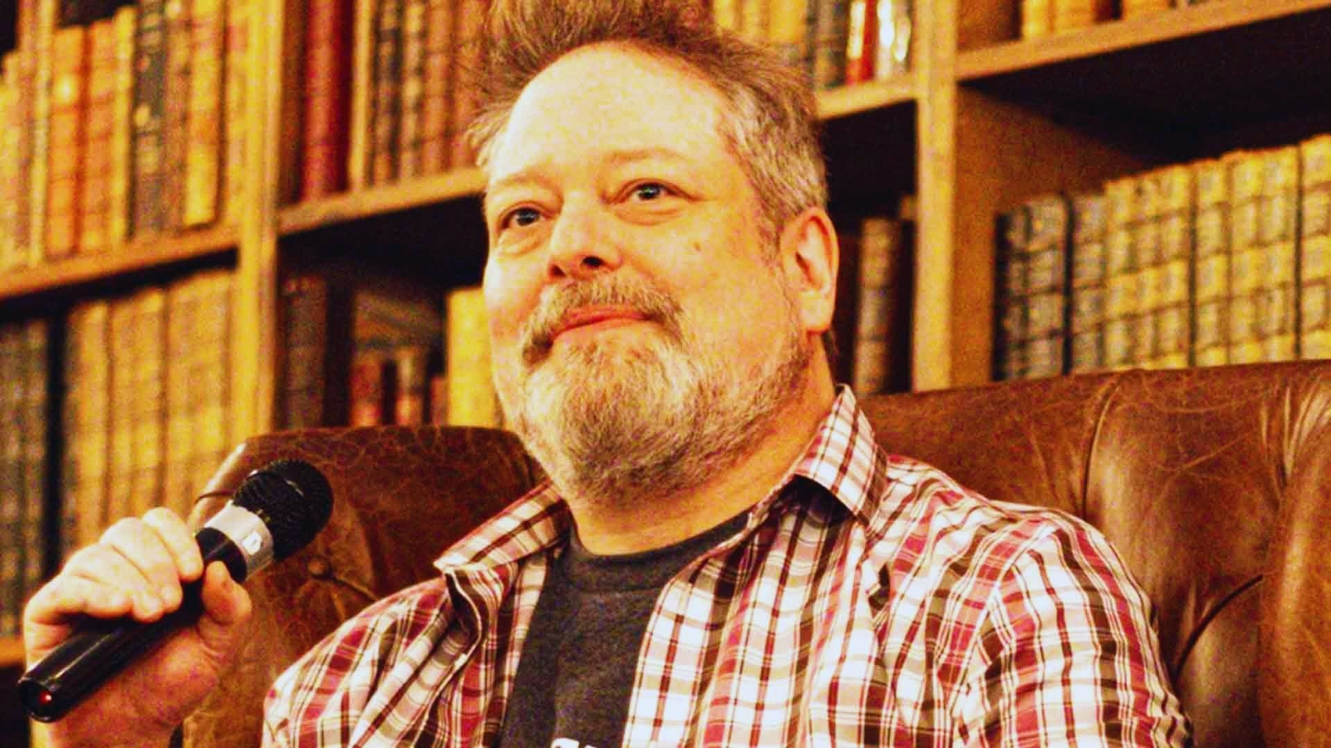 A bearded man sits in a leather chair, holding a microphone, with a thoughtful expression. Shelves filled with books form the cozy background.
