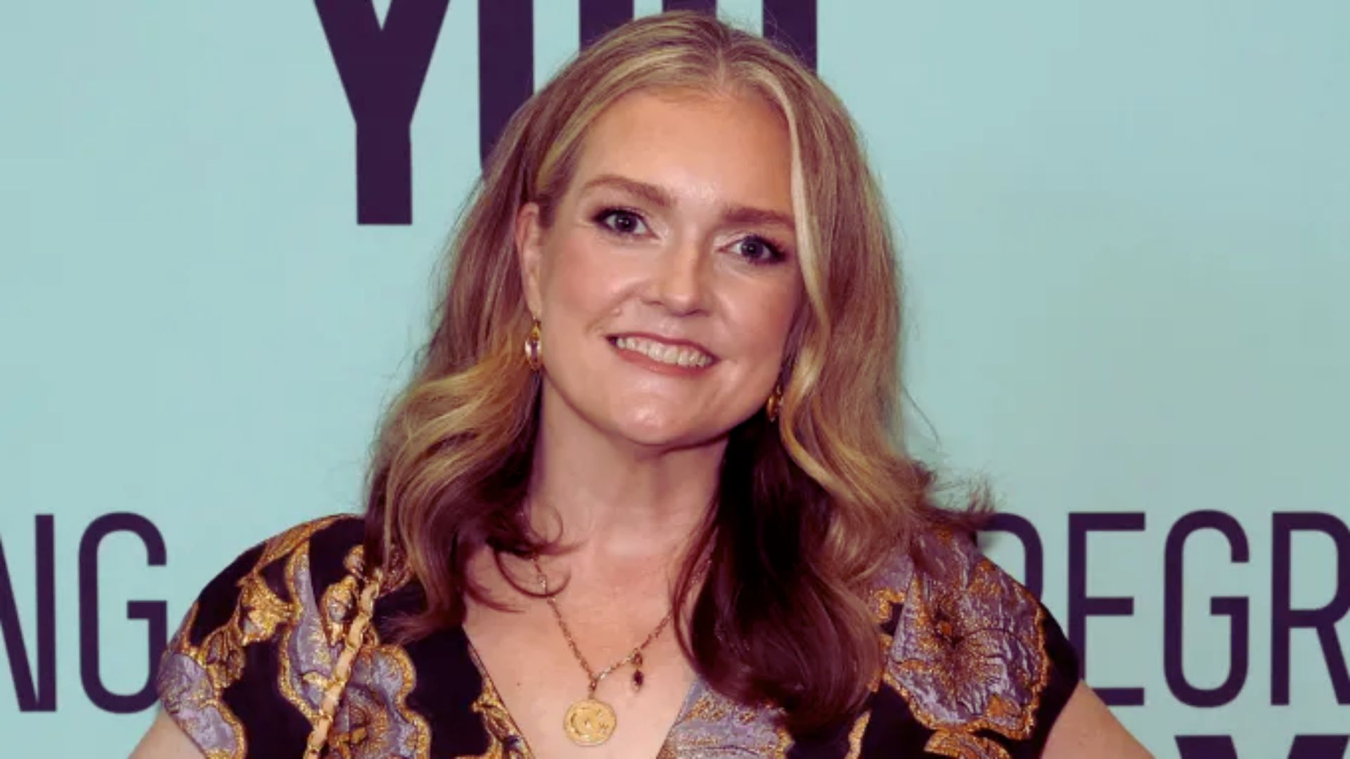 Woman with long, wavy, blonde hair wearing a patterned blouse and gold jewelry smiles warmly. Background features large text on a light blue wall.