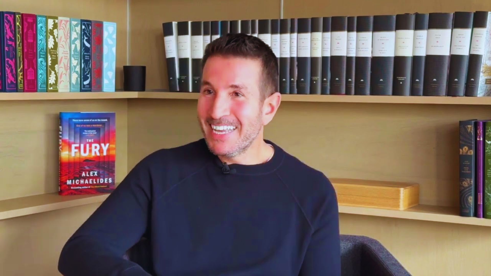 A person wearing a dark sweater is smiling while sitting in a cozy room with bookshelves. The shelves hold various colorful books and a prominently displayed book titled "Fury." The atmosphere is warm and inviting.
