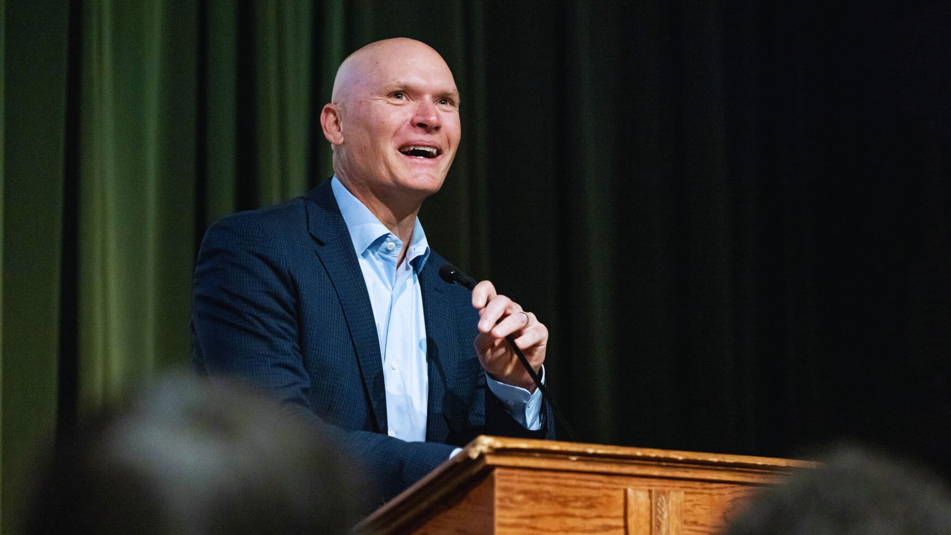 A bald man in a suit and tie delivers a speech at a podium during a formal event.