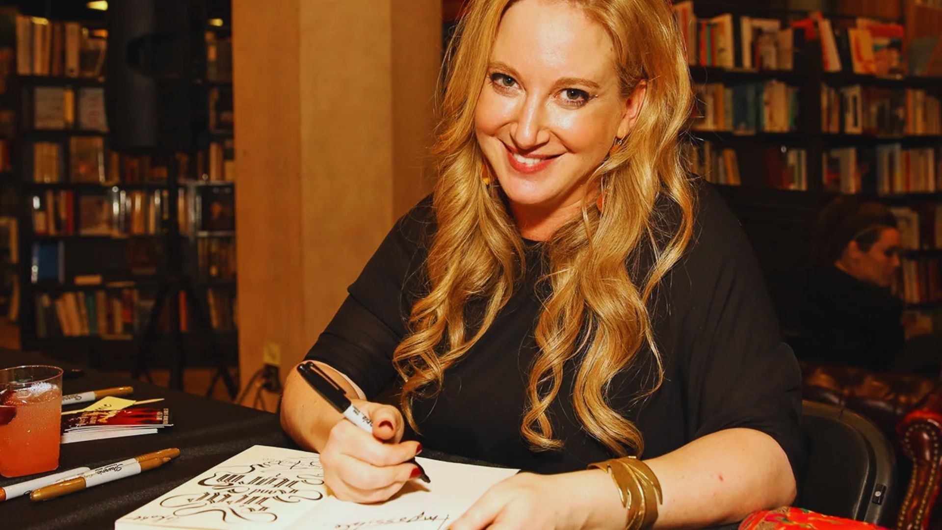 A smiling woman holds a pen, focused on writing on a notepad.