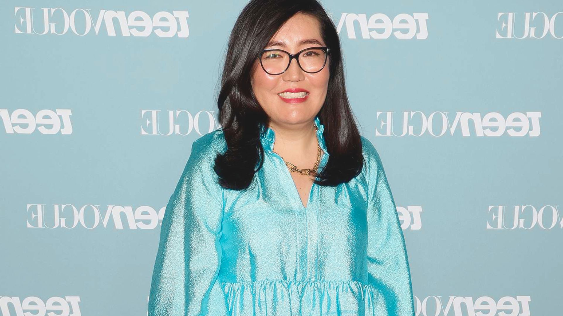 A woman wearing glasses and a blue dress stands confidently, smiling at the camera.