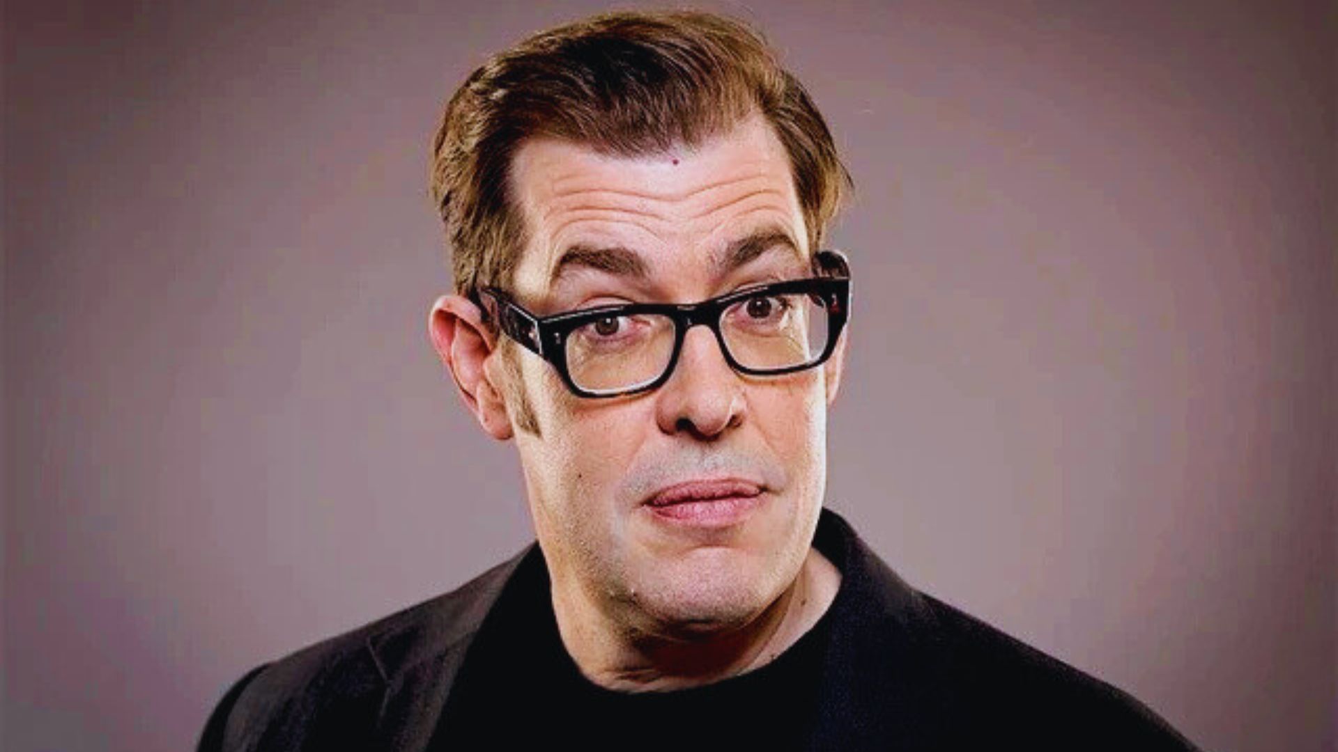A man wearing glasses and a black shirt, looking directly at the camera with a neutral expression.