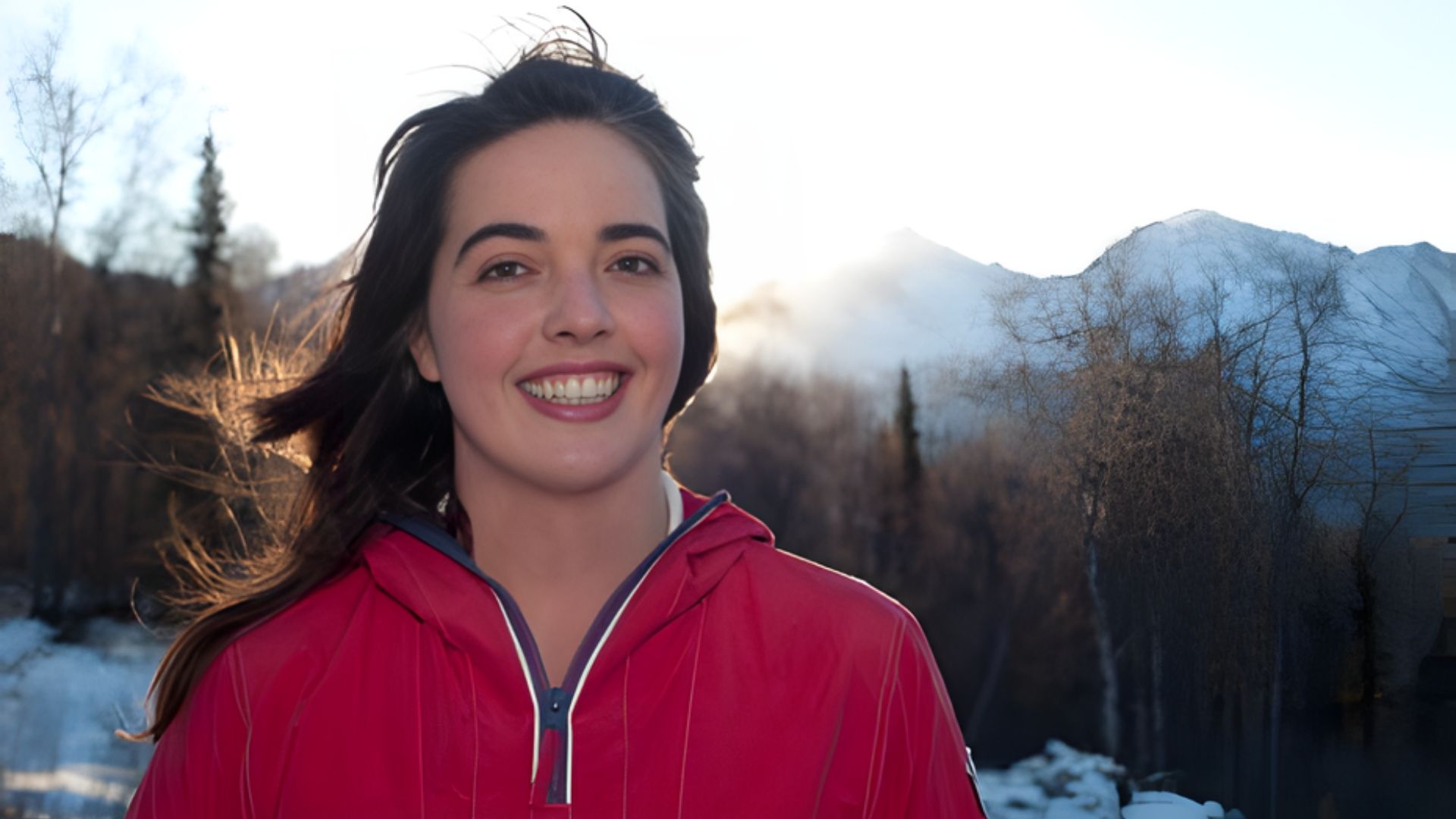 A woman wearing a red jacket smiles warmly at the camera, exuding a friendly and approachable demeanor.