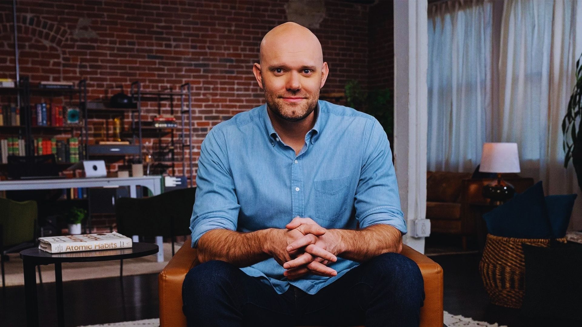 A bald man sits in a chair with his arms crossed, looking thoughtfully to the side.