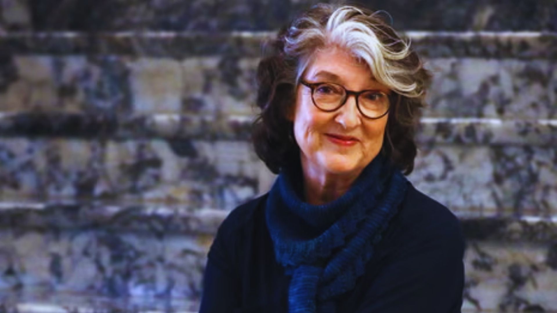 An older woman wearing glasses and a colorful scarf, smiling gently at the camera.