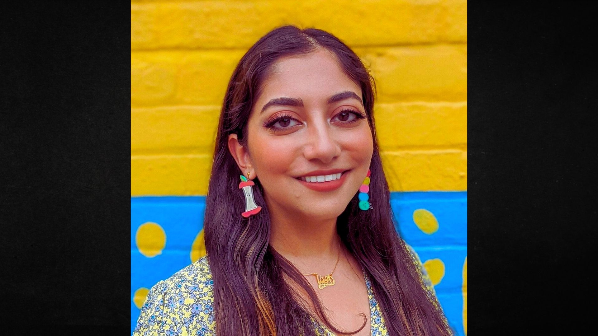 A woman with long hair and earrings stands in front of a bright yellow wall, smiling softly.