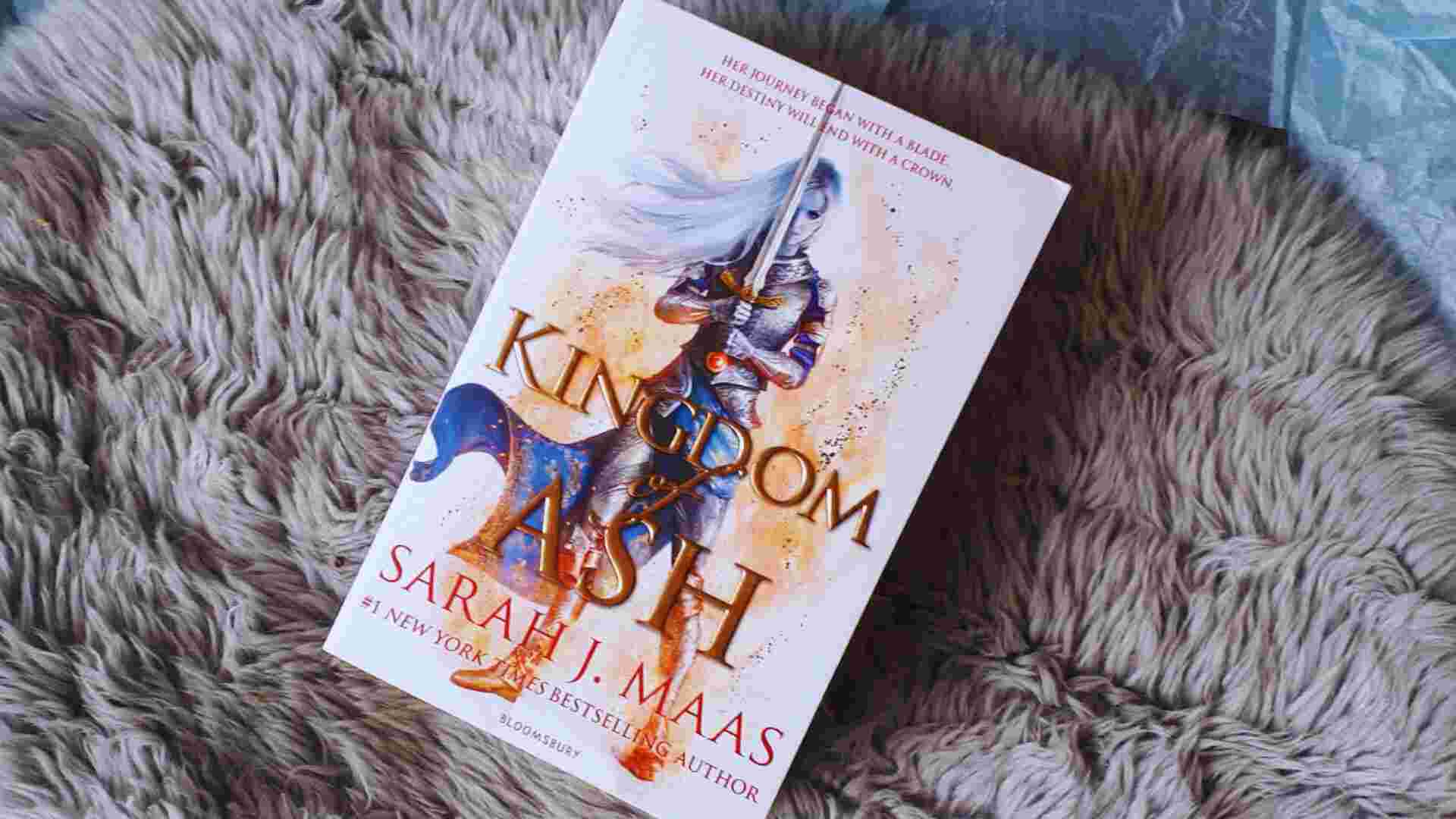 The book "Kingdom of Ash" by Sarah J. Maas lies on a shaggy fur rug. Its cover depicts a warrior woman with a sword, evoking a sense of strength and fantasy.