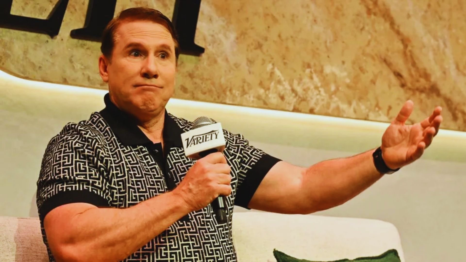 A person holding a "Variety" microphone gestures while speaking on stage. They wear a patterned shirt, appearing expressive against a textured backdrop.