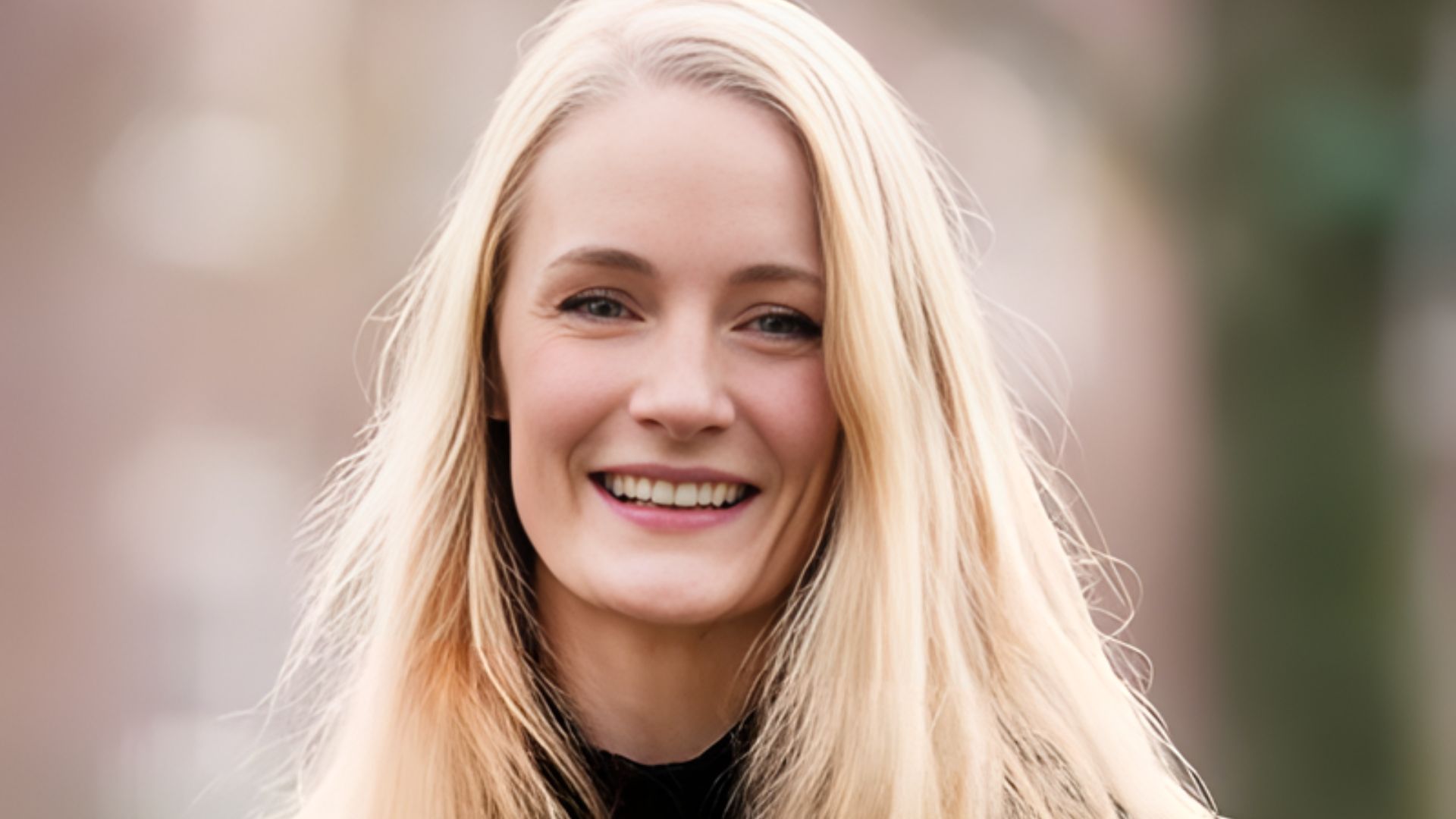 Smiling woman with long blonde hair stands outdoors, wearing a black top. The blurred background has soft, neutral tones, creating a warm, friendly atmosphere.