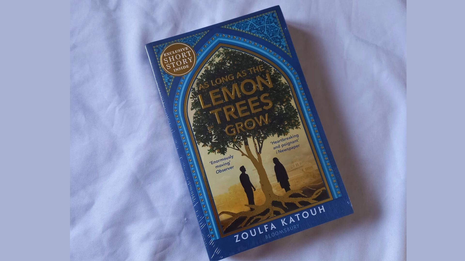 The book "Lemon Trees Grow" lies open on a neatly made bed, surrounded by soft pillows and a cozy blanket.