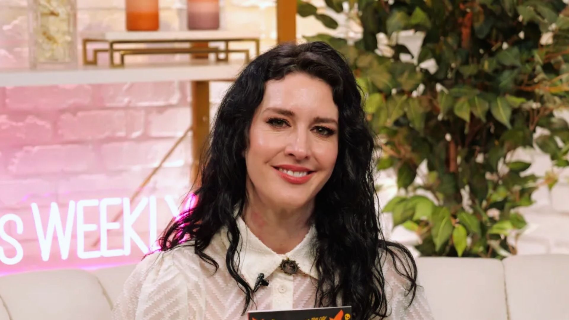 A woman with long black hair sits comfortably on a couch, looking relaxed and engaged in her surroundings.