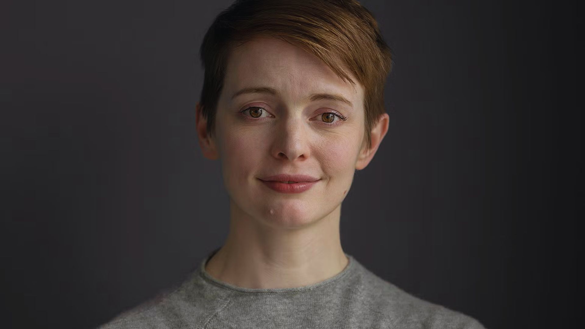 A woman with short hair wearing a grey shirt, looking confidently at the camera.