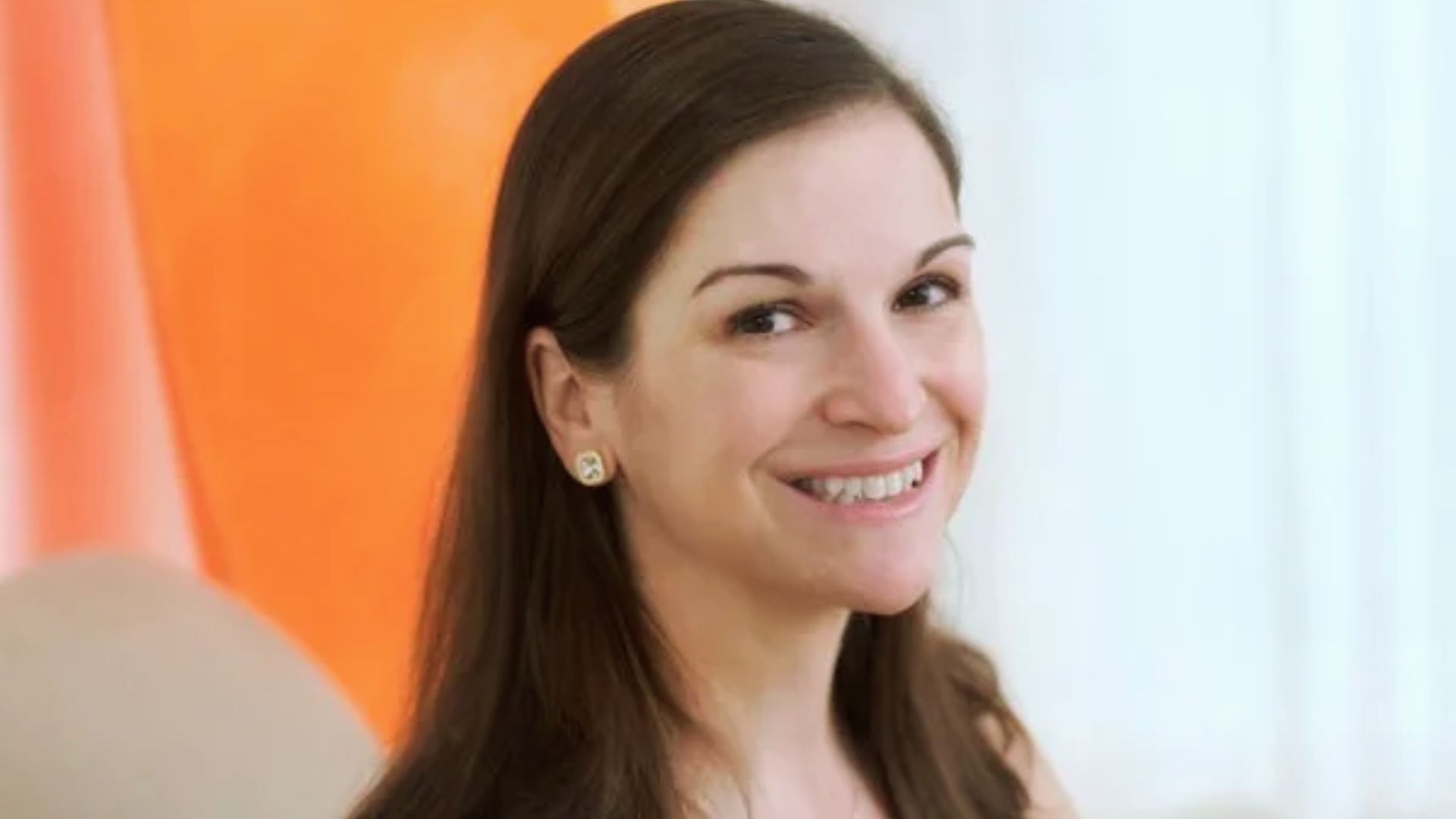 A woman smiling while seated in front of a vibrant orange curtain.