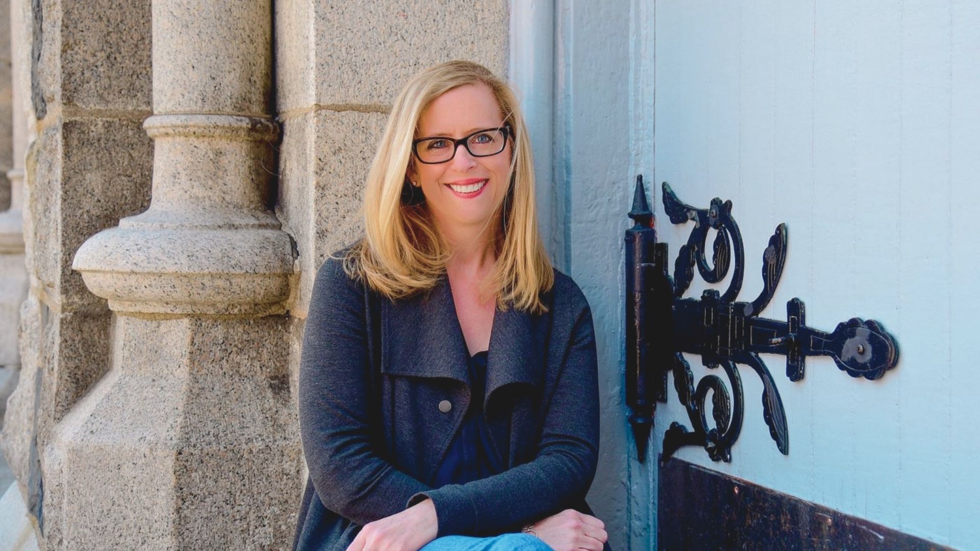 A woman wearing glasses sits on a bench, looking thoughtfully into the distance.
