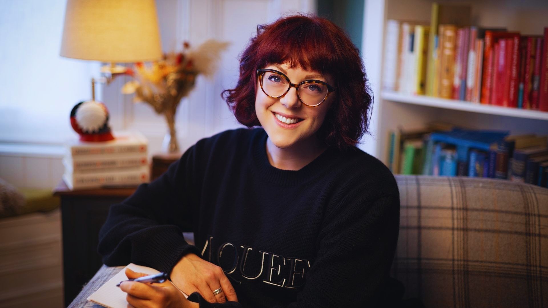 A woman with red hair and glasses sits on a couch, appearing thoughtful and at ease in her surroundings.