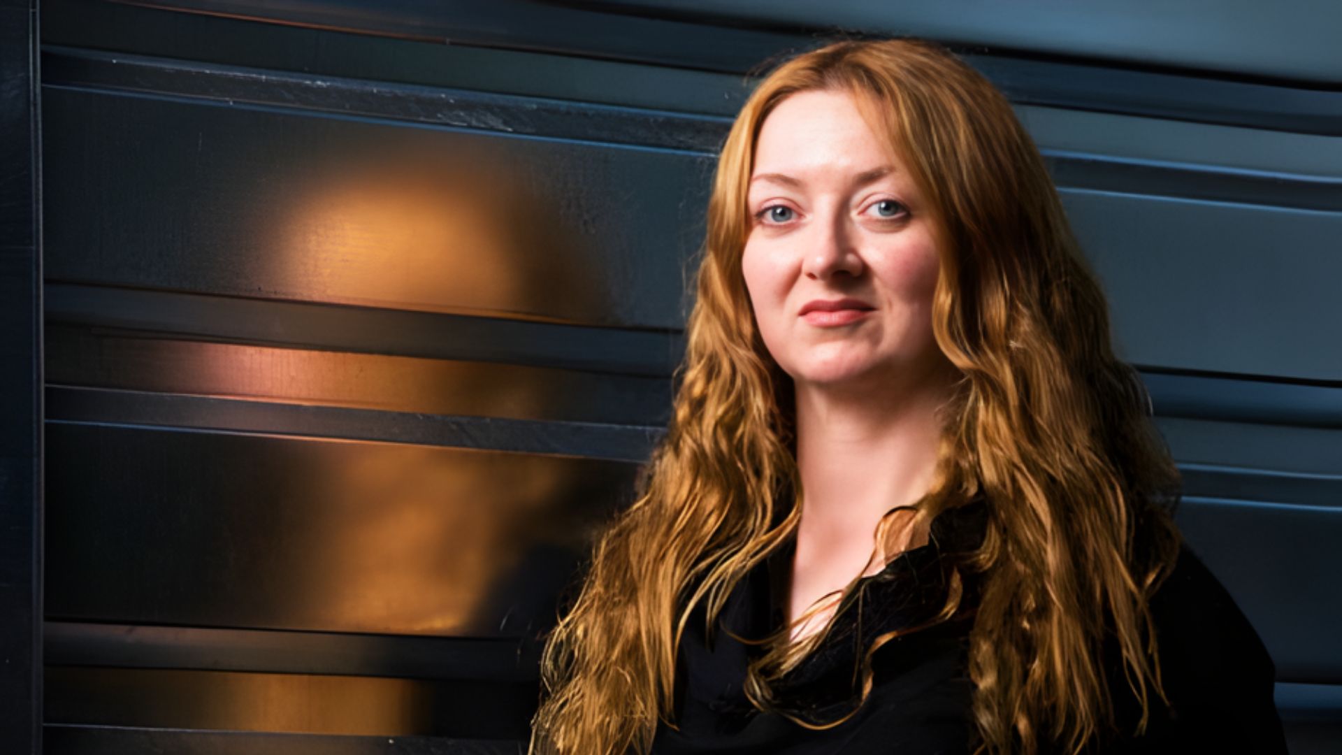 A woman with long red hair stands confidently in front of a large metal door.