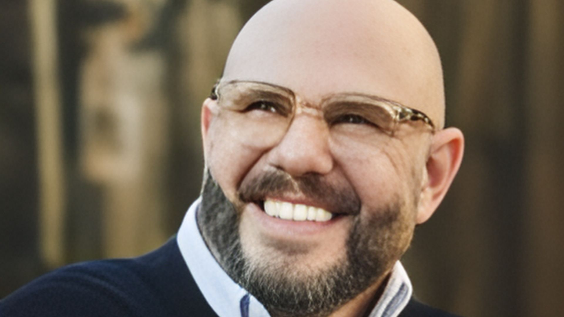 A bald man with glasses smiles while wearing a cozy sweater.