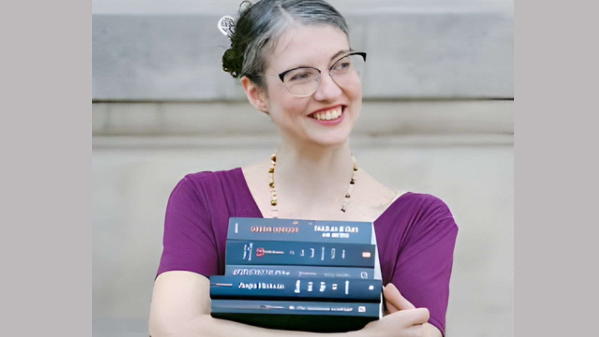 A woman with glasses holds a tall stack of books, smiling as she stands in a cozy reading nook.