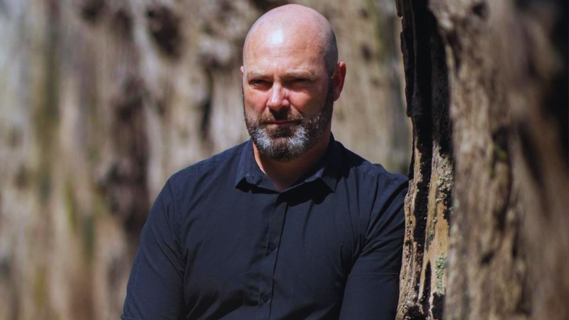 A bald man with a beard leans casually against a wall, looking off to the side.