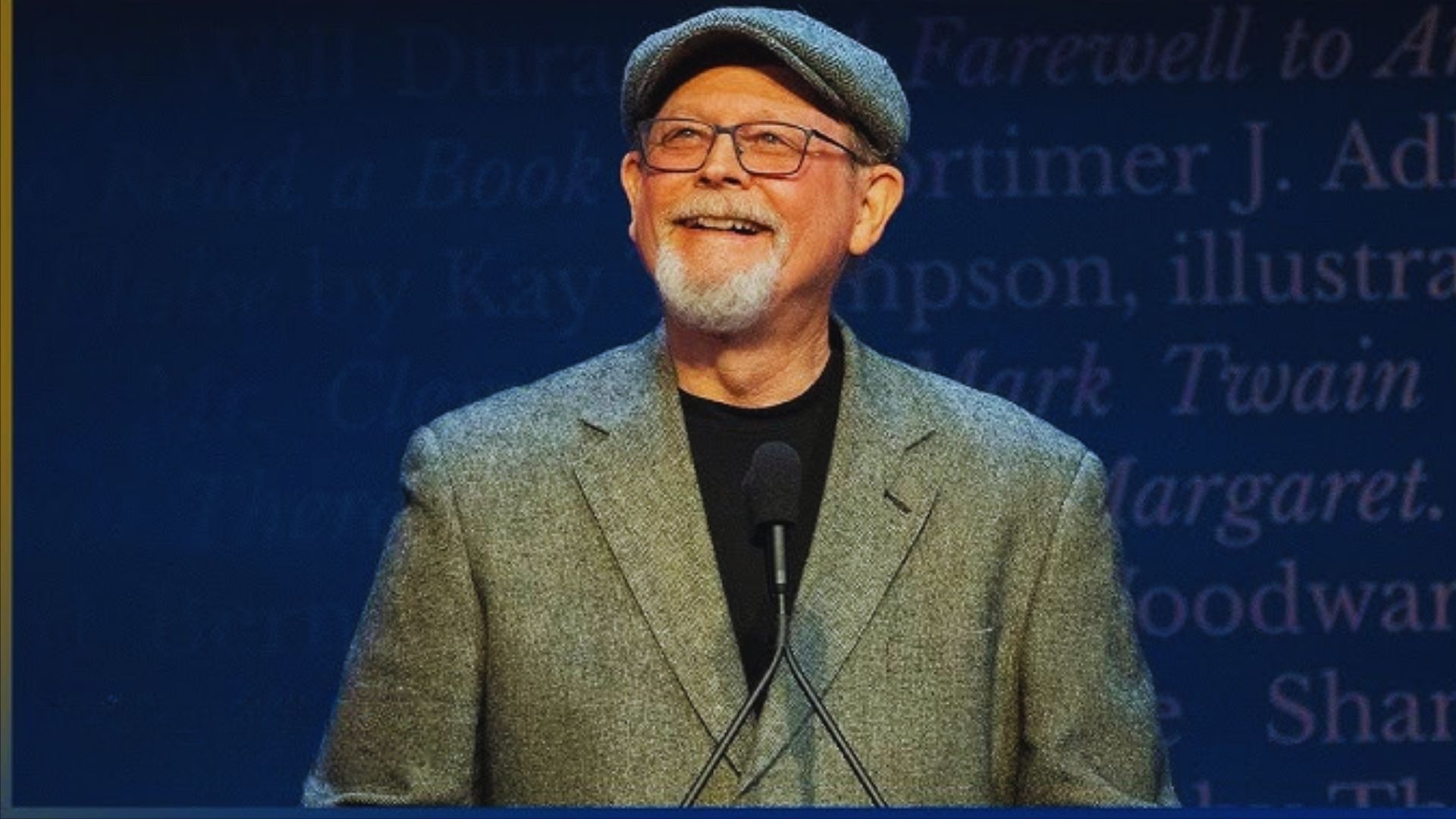 A man wearing a hat and glasses speaks at a podium, addressing an audience with a focused expression.