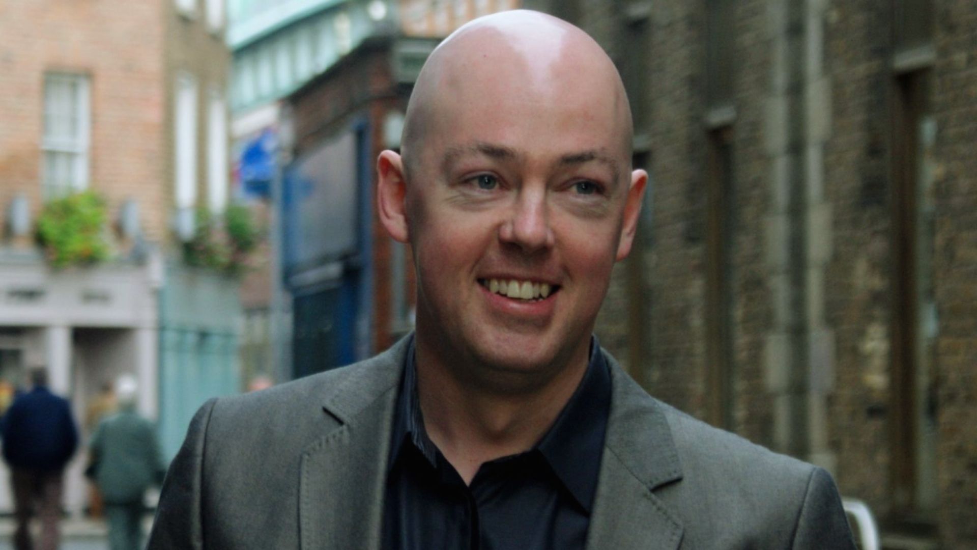A bald man in a suit smiles confidently while standing on a bustling city street.